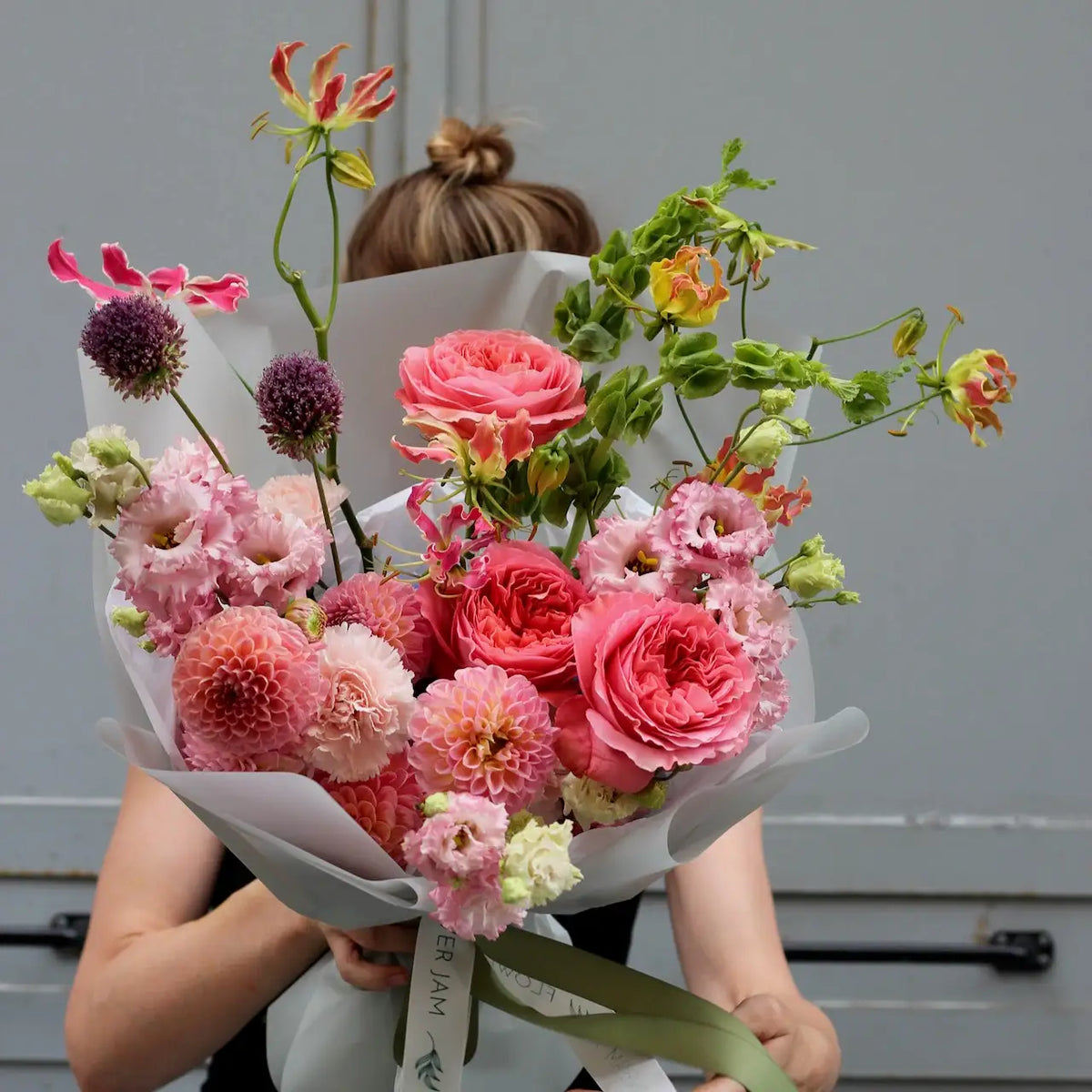 A woman holiding a bouquet of pink colored flowers on a grey background wrapped in a neutral paper.