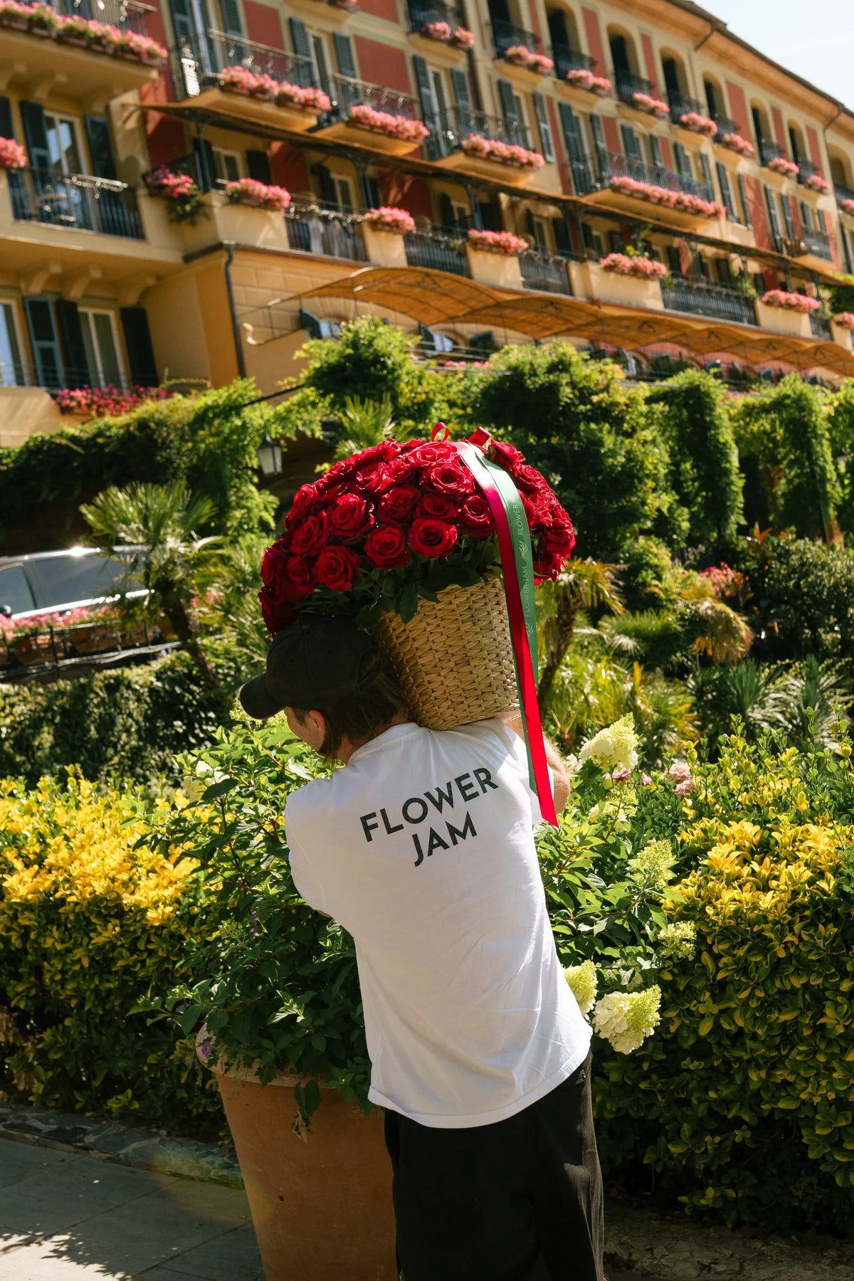 A person from behind wearing a white t-shirt with 'FLOWER JAM' printed on the back, carrying a large woven basket overflowing with vibrant red roses in a sunny garden setting with a hotel in the background.