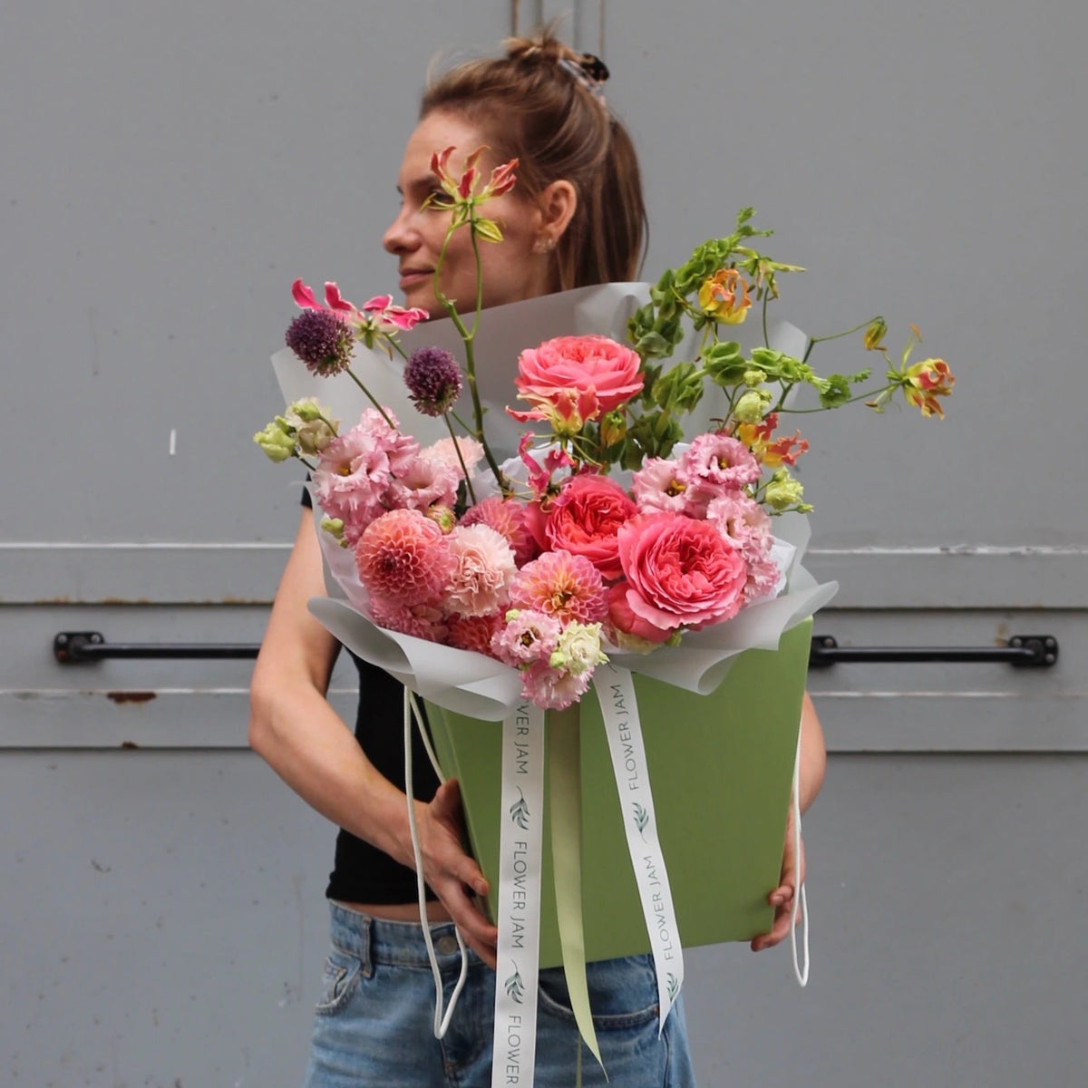 A woman holding a colorful bouquet of pink flowers inside a green holding bag against a grey background.