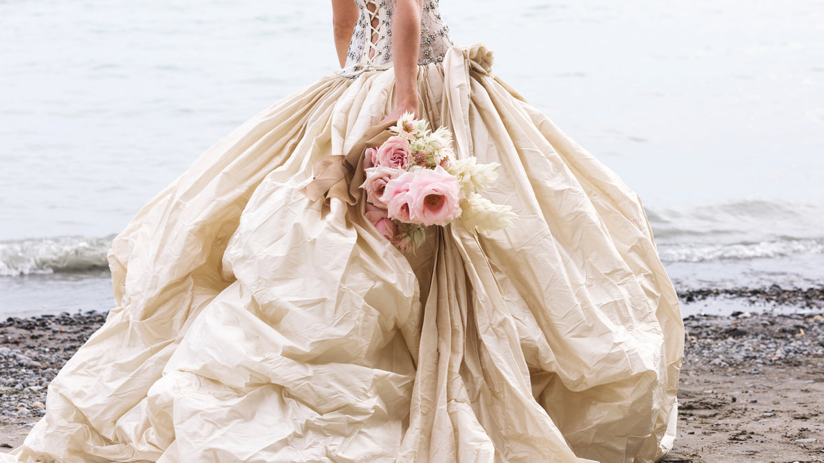 A woman in an ivory wedding gown, pacing on the beach and holding a bouquet of pink roses